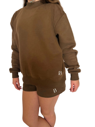 A person wearing an oversized brown crewneck sweatshirt and matching shorts against a solid pale pink background. A small white monogram logo 'B' is visible on both garments.